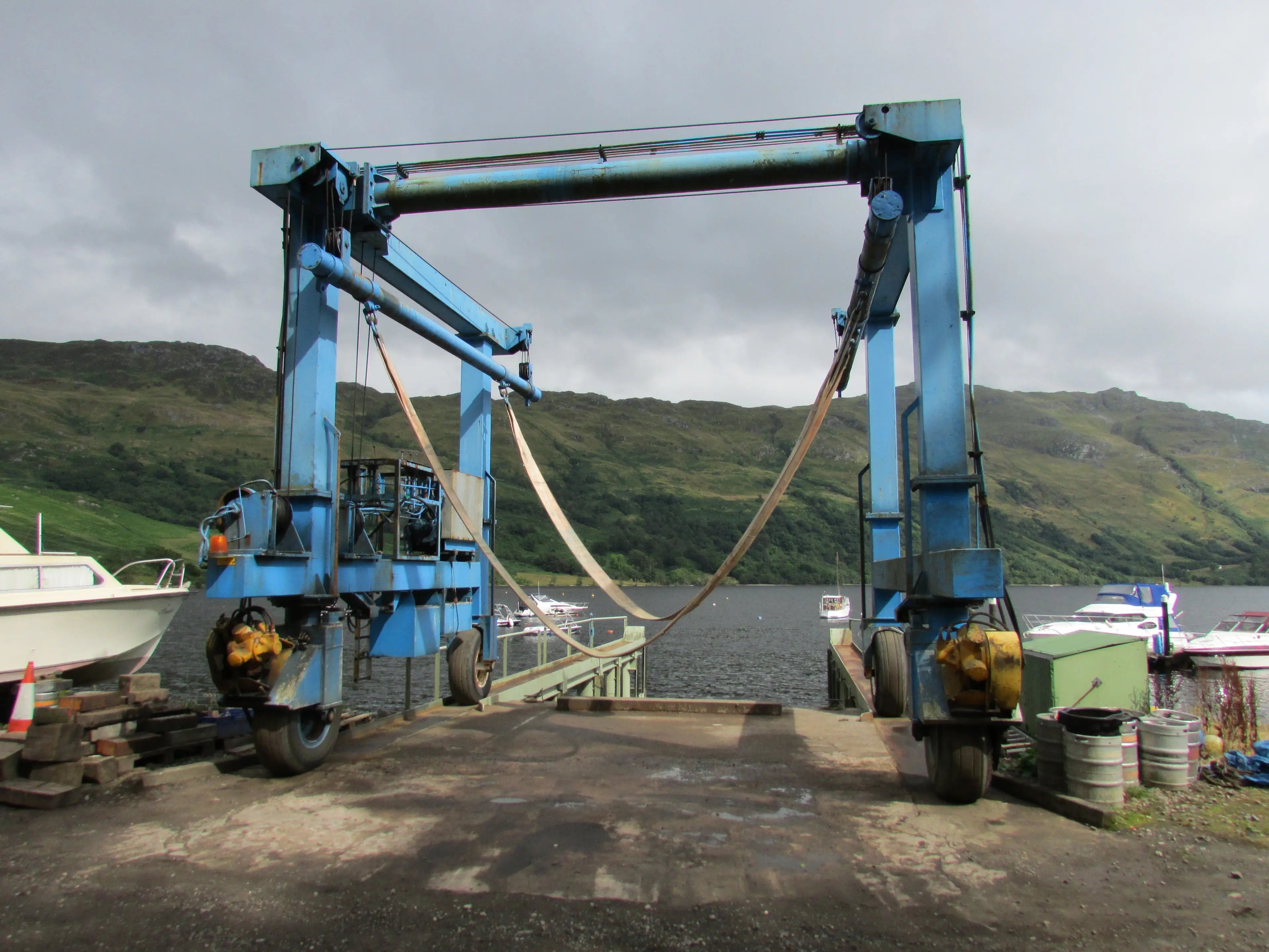 A boat hoist standing over a slipway with its lifting slings hanging empty, boats and a loch behind