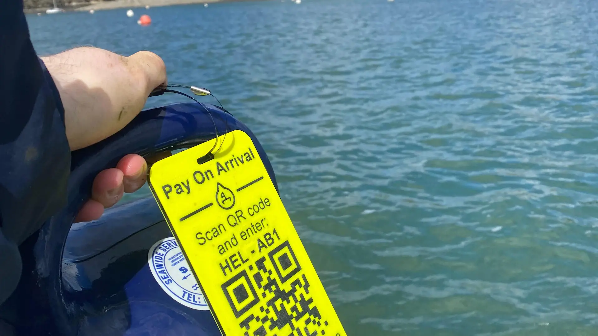 A yellow Pay On Arrival tag clipped to a mooring buoy, showing a QR code and the mooring's code, HEL-AB1